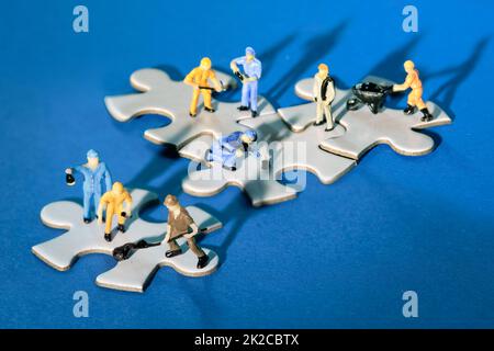 Squadra di minuscoli lavoratori in miniatura figure su jigsaw collegati puzzle pezzi isola su carta blu scuro. Luce spettacolare con ombre lunghe. Foto Stock