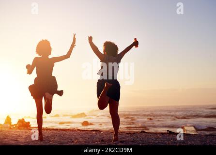 Amare la vita che viviamo insieme come amici. Ripresa da dietro di due giovani donne irriconoscibili che saltano nella gioia mentre camminano lungo la spiaggia al tramonto. Foto Stock