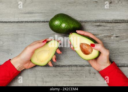 Vista del piano portapaziente, donna mano che regge due metà di avocado in ogni mano, ancora una pera intera collocato su grigio scrivania in legno sopra. Foto Stock