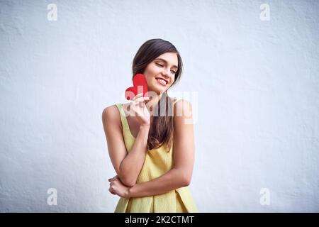 Gli shes hanno ottenuto l'amore sulla sua mente. Studio shot di una giovane donna allegra che tiene un pezzo di carta a forma di cuore nelle sue mani mentre si trova su uno sfondo grigio. Foto Stock