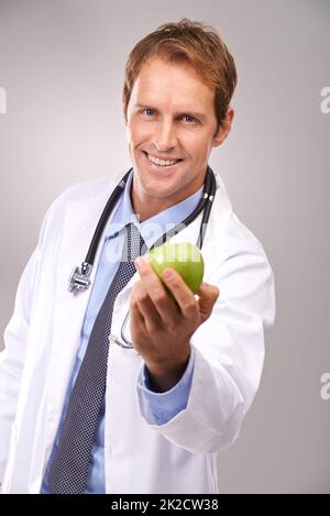 Uno al giorno mi terrà via. Studio ritratto di un bel giovane medico che tiene fuori una mela verde. Foto Stock