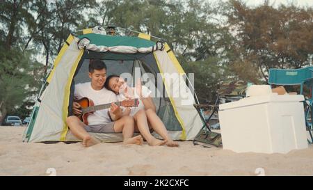 Giovane coppia amorevole grazioso mentre accampamento suona la chitarra mentre si siede alla tenda di campeggio sulla spiaggia in vacanza tempo al tramonto, giovane gruppo asiatico Foto Stock