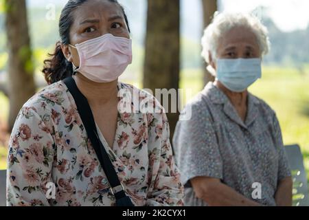 Asian anziano donna e figlia in sociale distanziamento seduta panca e indossare maschera viso per proteggere l'infezione di sicurezza Covid 19 Coronavirus in parco. Foto Stock