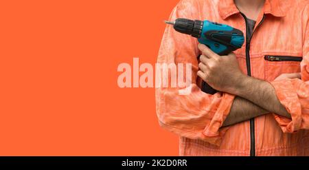 Capo meccanico che indossa un abito uniforme arancione che tiene il trapano elettrico blu di fronte a sfondo arancione. Ritratto con luce da studio. Foto Stock