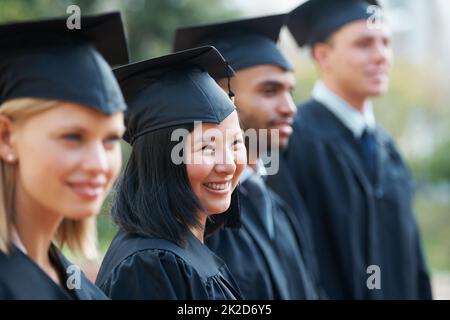 In attesa del loro futuro. Giovani laureati in possesso dei loro diplomi mentre si levano in piedi in fila e sorridenti. Foto Stock