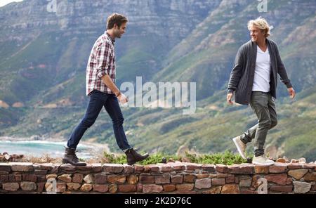 Collegamento con il mio amico all'aperto. Scatto di due giovani uomini che camminano su un muro di pietra con la montagna sullo sfondo. Foto Stock