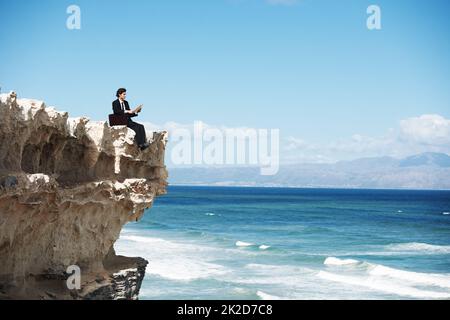 Liberarsi dalla corsa al ratto. Giovane uomo d'affari seduto sul bordo di una scogliera che si affaccia sull'oceano mentre legge un libro. Foto Stock