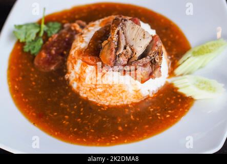 Carne di maiale alla griglia rossa su riso, cibo servito con curry dolce e verdure (cetriolo, coriandolo) su piastra bianca in ceramica. Foto Stock