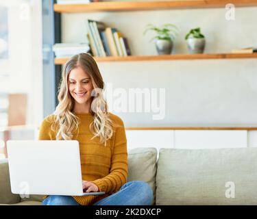 Aggiornare il suo blog. Scatto di una ragazza attraente usando il suo laptop mentre si siede sul divano a casa. Foto Stock