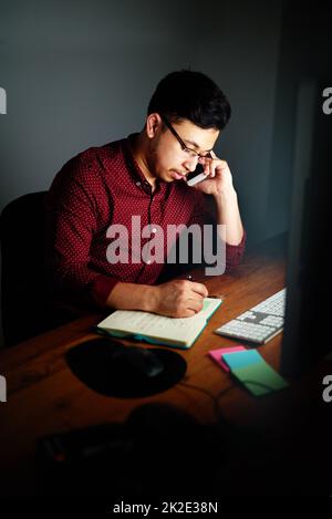 Mettendo in quelle ore di straordinario. Scatto di un giovane designer maschio che parla al telefono e prende appunti mentre lavora in ufficio a tarda notte. Foto Stock