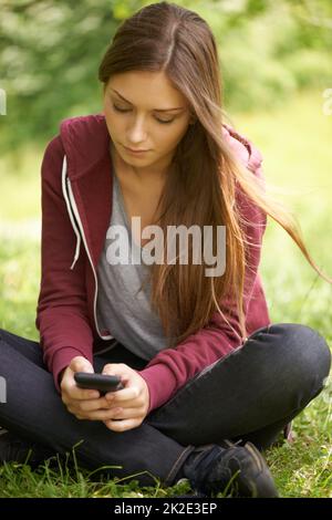 Sempre connesso. Una ragazza piuttosto giovane che texting sul suo telefono mentre si siede in un campo verde. Foto Stock