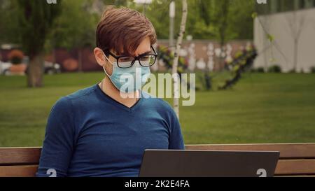 L'uomo lavora sul suo computer portatile mentre era seduto su una panca all'aperto in slow motion. Espressioni facciali focalizzate con maschera chirurgica. Lavorare in modo responsabile. Lavorare durante la pandemia. Foto Stock