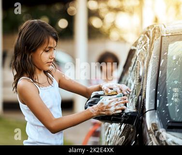 Ottenere la macchina bella e pulita. Scatto di una ragazza piccola allegra lavando i suoi genitori auto fuori durante il giorno. Foto Stock