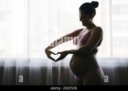 Il suo cuore sta crescendo insieme al suo bambino. Scatto corto di una giovane donna incinta attraente in piedi nella sua camera da letto a casa. Foto Stock