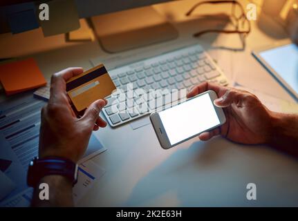 Ordinare il take-out per fargli passare la notte. Ripresa ritagliata di un uomo d'affari irriconoscibile utilizzando una carta di credito e un telefono cellulare durante una tarda notte al lavoro. Foto Stock