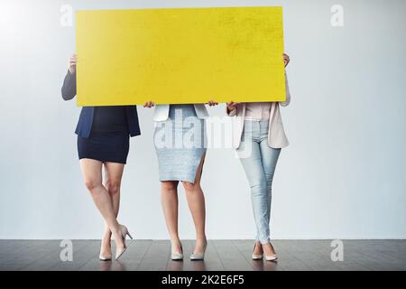 Ottenga dietro questo marchio di affari. Studio girato di un gruppo di donne d'affari che tengono su un cartello giallo bianco davanti a loro. Foto Stock