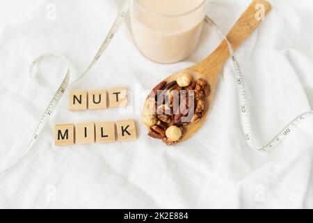 Latte vegano di noci in una tazza di vetro con vari dadi su un tavolo bianco, noci in un cucchiaio di legno. Latte di noci con scritte. Foto Stock