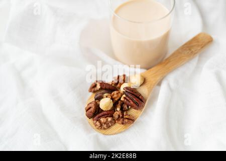 Latte vegano di noci in una tazza di vetro con vari dadi su un tavolo bianco, noci in un cucchiaio di legno. Foto Stock