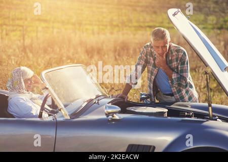 Problemi con l'auto RoadTrip. Colpo di un uomo anziano che guarda sotto il cofano della sua auto durante un viaggio con sua moglie. Foto Stock