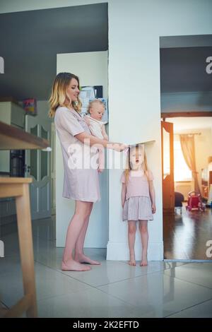 Sono cresciuto più in alto durante la notte. Colpo di una madre che porta il suo bambino e che misura l'altezza delle sue figlie contro un muro. Foto Stock