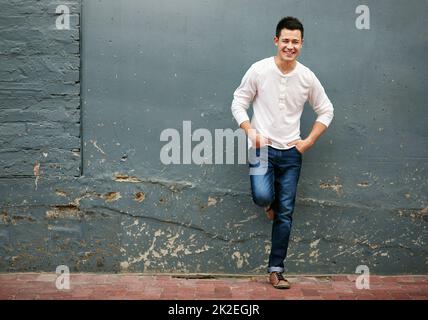 HES un ragazzo facile-andare. Ritratto di un bel giovane uomo in piedi con le mani in tasca contro un muro grigio. Foto Stock