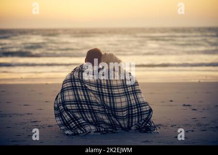 Theres niente più romantico che guardando il tramonto. Scatto retroguarato di una giovane coppia seduta sulla spiaggia con una coperta avvolta intorno loro. Foto Stock