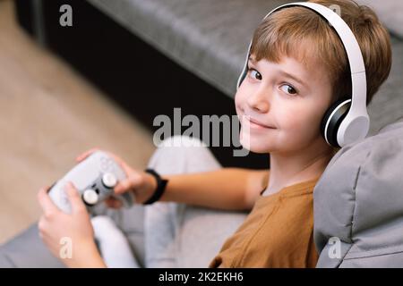 Adolescente soddisfatto in cuffie e con il joystick nelle sue mani guarda la fotocamera mentre si siede sulla sedia del sacco del fagiolo Foto Stock