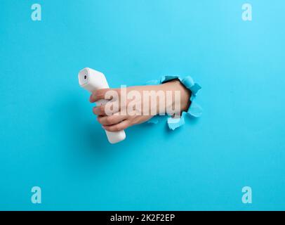 una mano femmina tiene un termometro in plastica senza contatto per misurare la temperatura di una persona, una parte del corpo sporge da un foro lacerato su uno sfondo di carta blu Foto Stock
