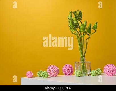 vaso trasparente in vetro con fiori secchi verdi su un tavolo bianco, sfondo giallo Foto Stock