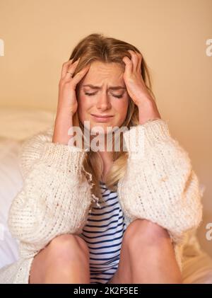 Il dolore è quasi insopportabile. Scatto di una giovane donna con un mal di testa che tiene la testa mentre si siede a letto. Foto Stock