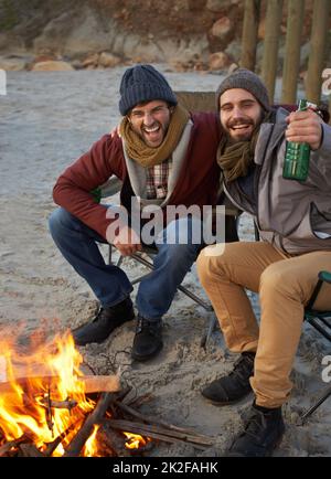 Riscaldarsi con un fuoco e una birra. Due giovani uomini seduti intorno ad un fuoco sulla spiaggia. Foto Stock