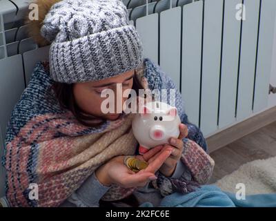 Donna che conta monete in euro e tiene il piggy bank vicino alla batteria a casa. Donna vestita calorosamente è sconvolta circa il prezzo elevato di riscaldamento. Costoso it Foto Stock