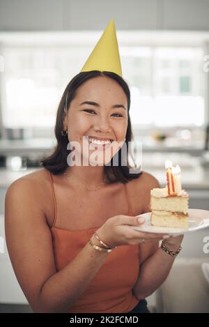 Chi non ama la torta di compleanno. Scatto di una giovane donna in posa con un pezzo di torta a casa. Foto Stock