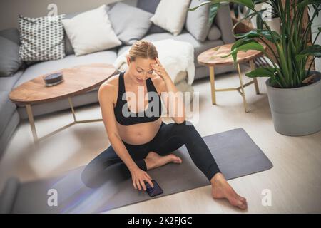 Gravidanza e tecnoloy supportato sano stile di vita concetto. Allegro felice donna incinta utilizzando l'applicazione smartphone mentre si esercita sul tappetino yoga sul pavimento del soggiorno a casa. Foto Stock