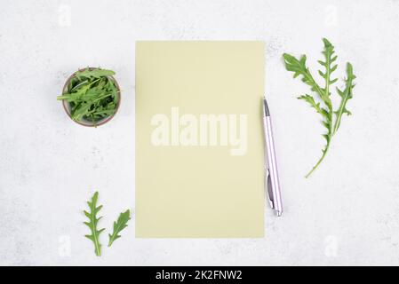 Carta bianca colorata con penna, foglie di insalata di rucola, spazio per la copia del testo, ricetta per cucinare, preparazione del cibo, stile di vita sano, dieta Foto Stock