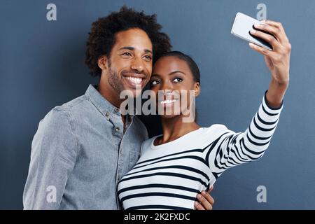 Nuova immagine del profilo. Scatto di una giovane coppia felice scattando una foto di se stessi con un cellulare su sfondo grigio. Foto Stock