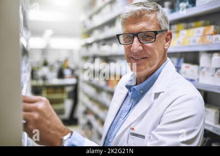 Immagazzinare su alcuni nuovi meds. Ritratto di un farmacista maschio maturo allegro ottenere farmaci da uno scaffale mentre si guarda la macchina fotografica in una farmacia. Foto Stock