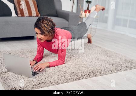 Il multitasking è la mia specialità. Scatto a tutta lunghezza di una giovane e attraente donna che fa il multitasking e che fa pilates mentre usando un laptop nel salotto. Foto Stock