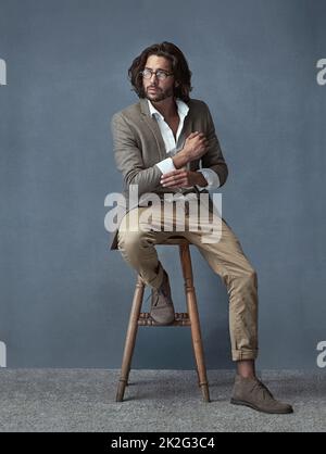 HES ha lo stile. Scatto completo in studio di un bel giovane uomo seduto su uno sgabello su sfondo grigio. Foto Stock