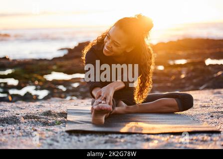 Il mio corpo mi ringrazia per ogni pratica di yoga. Scatto di una giovane donna attraente seduta su un tappeto e stretching mentre si fa yoga sulla spiaggia al tramonto. Foto Stock