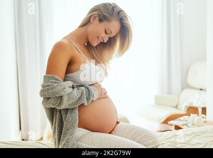 Non vedo l'ora di tenerti tra le mie braccia. Shot di una donna incinta che tocca il ventre mentre si siede sul suo letto. Foto Stock