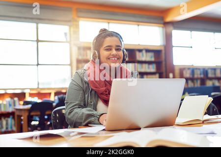 La musica fa la sua esecuzione. Ritratto di una giovane e allegra studentessa che lavora su un computer portatile mentre ascolta musica con le sue cuffie all'interno di una biblioteca. Foto Stock