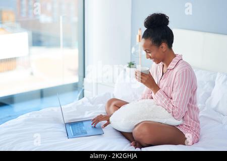 Passando attraverso il mio blog prima che la mia giornata comincia. Scatto di una giovane donna attraente bere caffè mentre usando il suo portatile a letto a casa. Foto Stock