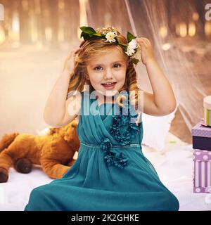 I fiori sono importanti per una principessa di fata. Scatto di una ragazza felice che guarda la macchina fotografica e tocca una corona di fiori sulla sua testa mentre si siede fuori nel bosco. Foto Stock