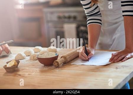 Si impara facendo. Scatto ritagliato di una donna che scrive una ricetta per l'impasto che prepara la pasta. Foto Stock