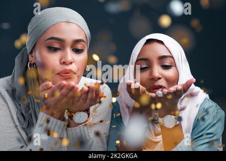 Lascia un po' di scintilla ovunque vai. Scatto corto di due giovani donne attraenti che indossano hijabs e che soffiano i coriandoli mentre si levano in piedi contro uno sfondo nero dello studio. Foto Stock