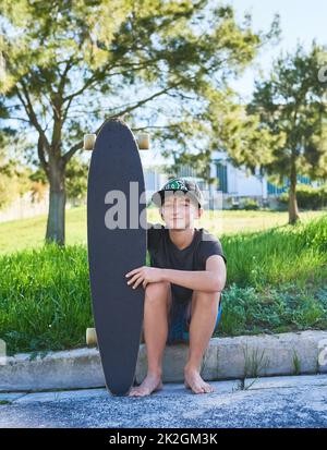 Imparare a pattinare. Ritratto a tutta lunghezza di un ragazzo seduto sul marciapiede con la sua longboard. Foto Stock