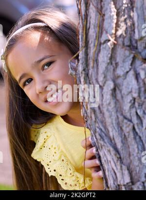 Non raccontare a nessuno il mio luogo preferito di nascondiglio e ricerca. Scatto di una bambina adorabile che si nasconde dietro un albero. Foto Stock