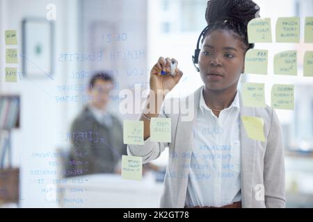 Prendendo nota delle principali preoccupazioni dei suoi clienti. Scatto di una giovane donna d'affari che indossa una cuffia mentre si brainstorming con note su una parete di vetro in un ufficio. Foto Stock