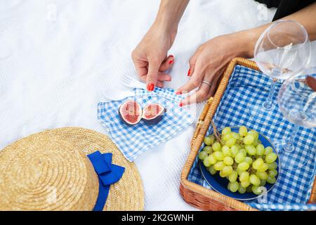 Romantico pic-nic nel parco sull'erba, cibo delizioso: Cesto, vino, uva, fichi, formaggio, tovaglia a scacchi blu, due bicchieri di vino. Ragazza taglia fichi.Outdoor concetto di ricreazione. Foto Stock